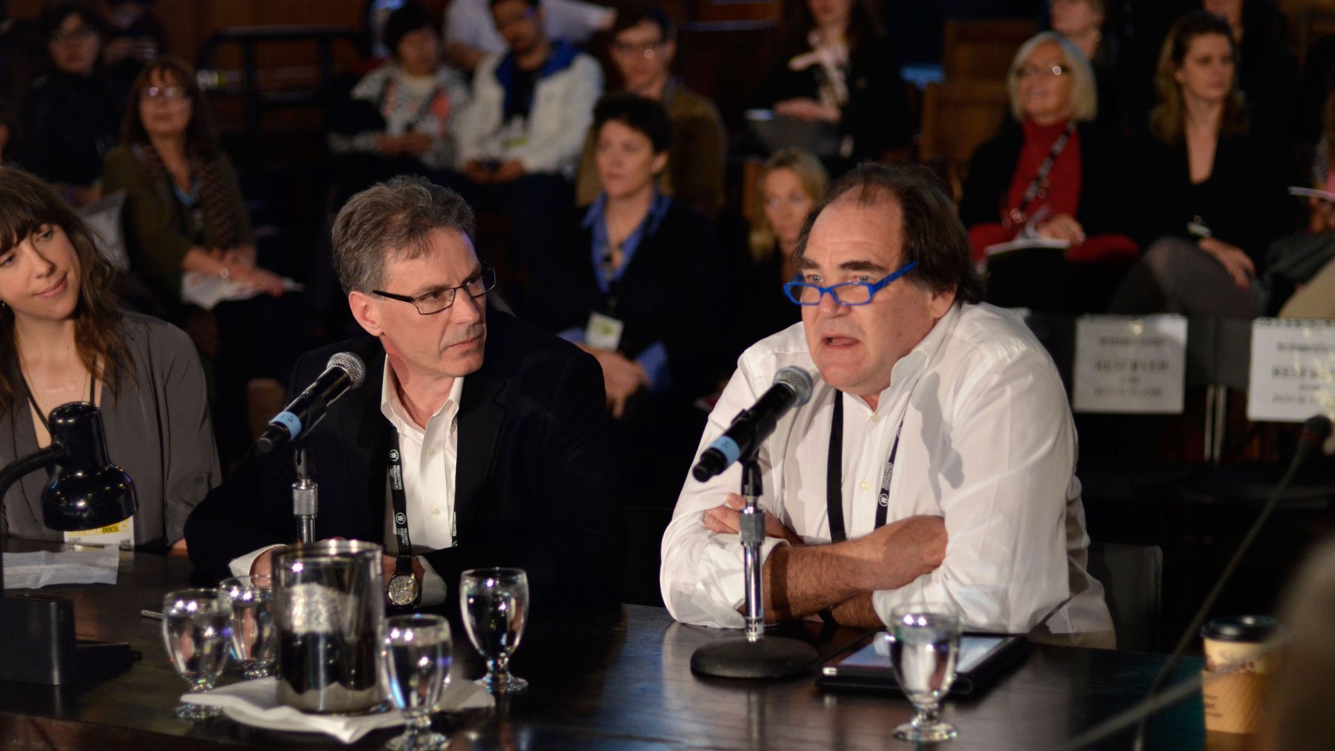 Three people sit at a table with microphones in front of them, one speaking into the microphone in an indoor theatre filled with audience members behind the speakers.