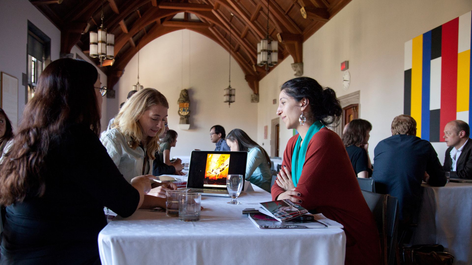 People in conversation seated at a table with white table cloth and looking at a laptop screen indoors.